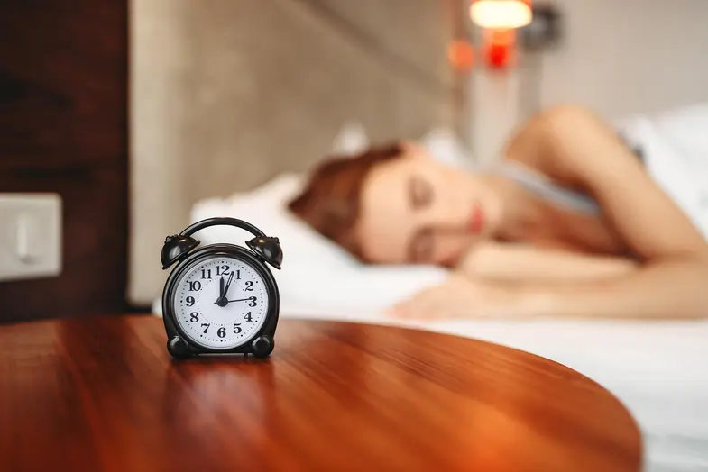 Woman sleeping next to a bedside table with an alarm clock on it | Justbob