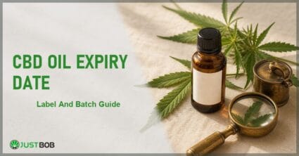 Justbob CBD Oil Expiry Date banner with green title, amber CBD oil bottle with blank label, hemp leaf and brass loupe on cream linen