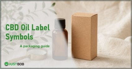 Justbob CBD Oil Label Symbols banner with green title, a CBD oil bottle, a small kraft box and a brass loupe on cream linen