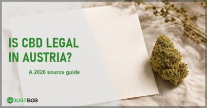 Justbob Is CBD Legal In Austria banner with green title, a hemp flower bud and a blank document sheet on cream linen