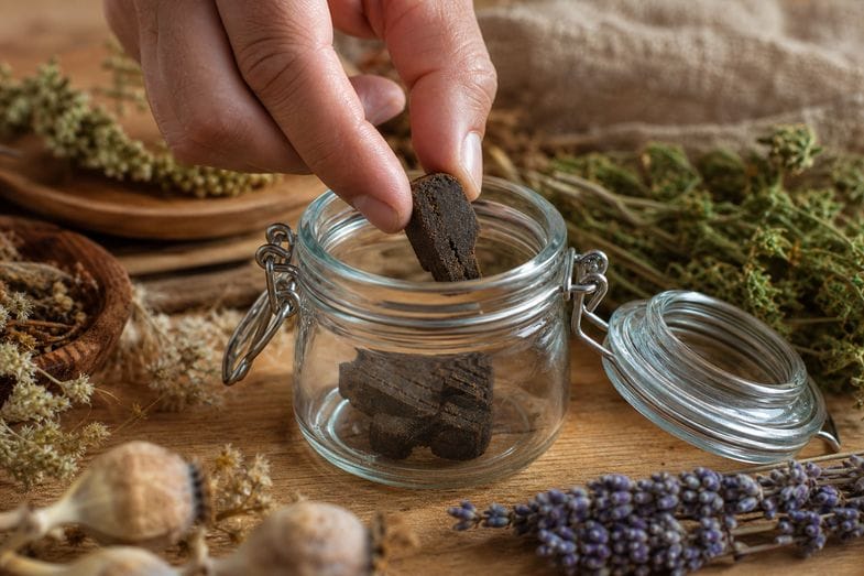 A hand placing a piece of dark CBD hash into a clear glass preserving jar with dried botanicals
