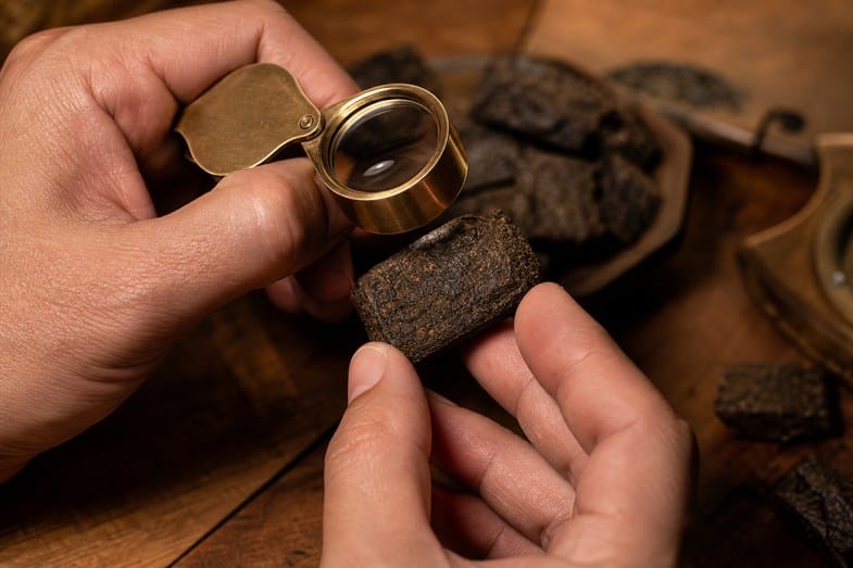 old-cannabis-and-old-hash-documents Hands inspecting a piece of aged pressed CBD hash under a brass loupe on a wooden table