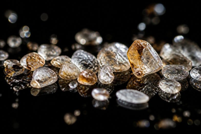 cbd-vs-cbda-crystal-macro Translucent white and amber crystal fragments scattered on a dark reflective surface and lit from behind